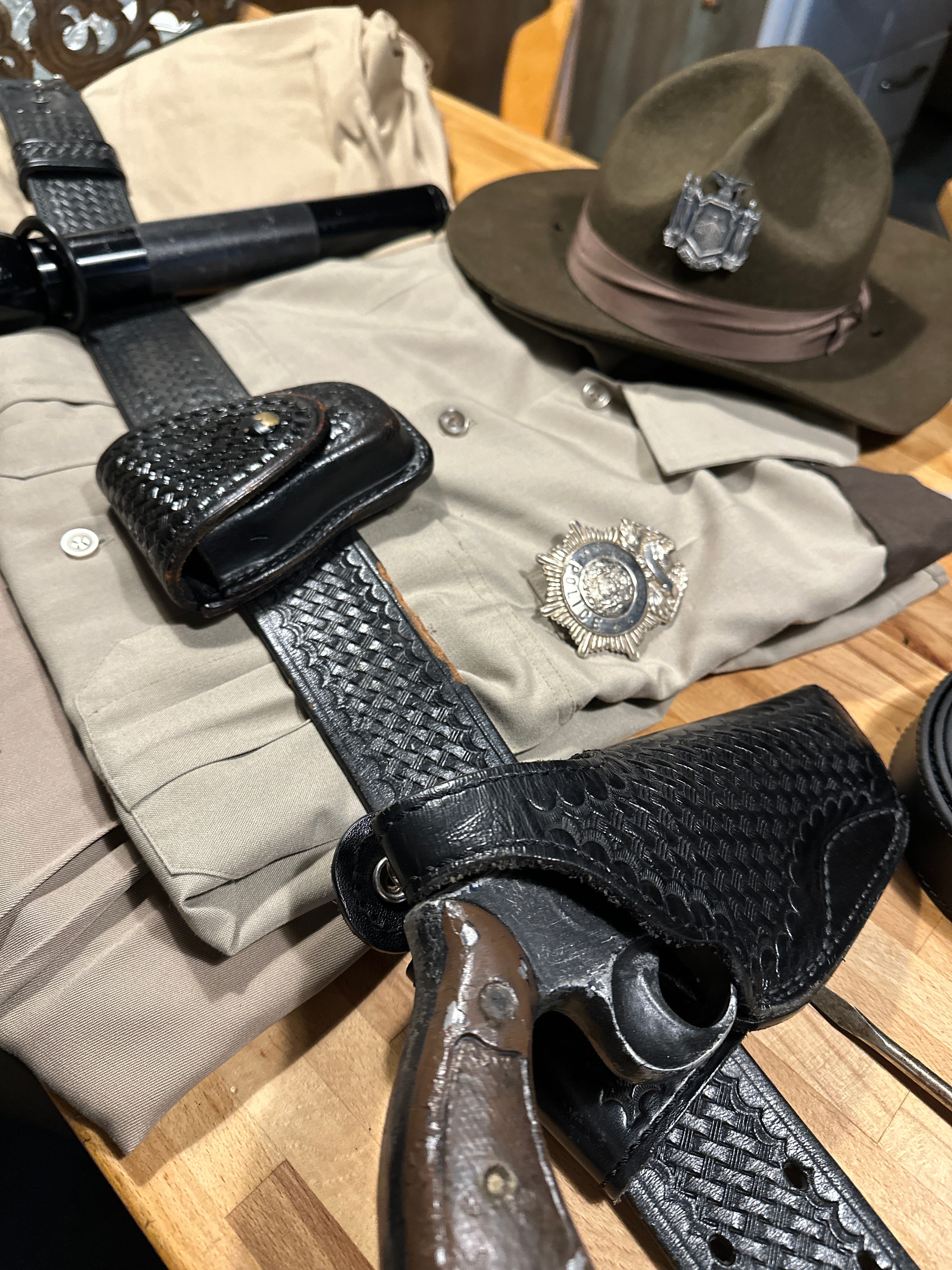 Vintage country police accessories — olive campaign hat, tan uniform shirt with badge, black basket-weave leather duty belt with holster and baton ring